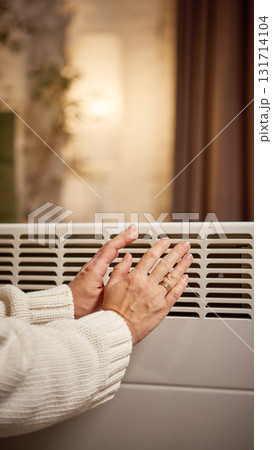 Hands warming near electric convector in softly lit living room 131714104