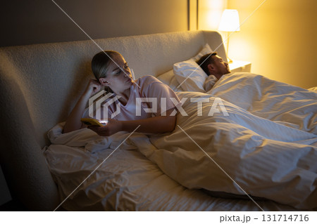 Woman using smartphone in bed secretly messaging someone while her partner sleeps beside her Woman using smartphone in bed secretly messaging someone while her partner sleeps beside her 131714716