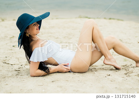 A girl in a white swimsuit is lying on the warm sand, adorned with a blue hat A girl in a white swimsuit is lying on the warm sand, adorned with a blue hat 131715140