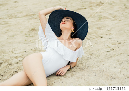 A young woman in a white swimsuit is sitting on the beach with a blue hat A young woman in a white swimsuit is sitting on the beach with a blue hat 131715151