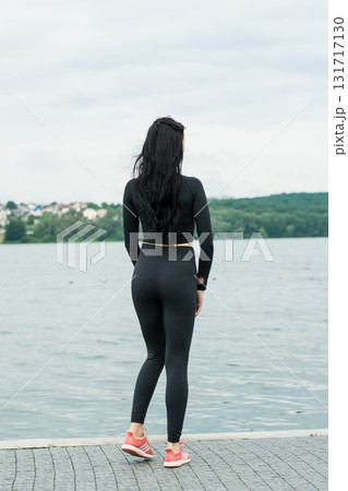 A girl wearing black leggings and a tight sweatshirt is performing gymnastic exercises on the shore of a lake 131717130