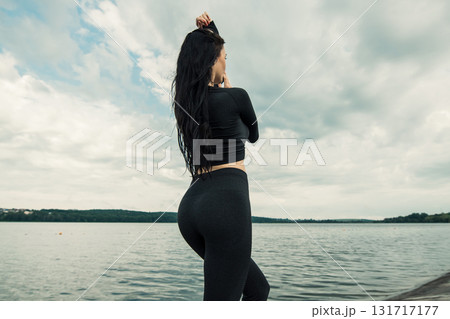 A young girl with an athletic build stands against the backdrop of a lake in black leggings and a sweatshirt 131717177