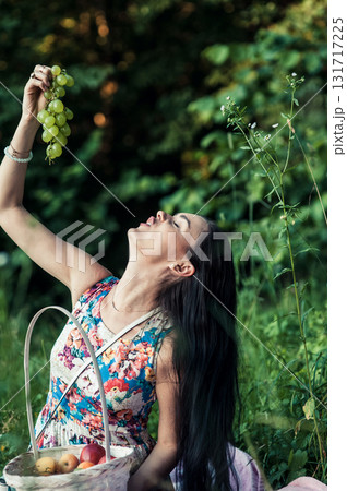 A girl at a picnic in the park holds a bunch of grapes against a backdrop of green trees 131717225