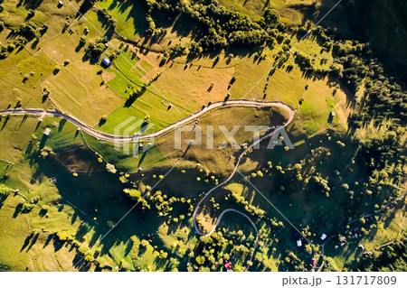 Top-down aerial view of picturesque rural landscape bathed in golden sunlight. Winding roads meander through green fields and small patches of trees. Scattered farmhouses and haystacks dot terrain. 131717809