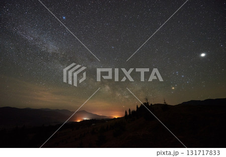 Breathtaking night sky displays dense field of stars and Milky Way over tranquil mountain landscape. Silhouetted trees and distant mountain peaks faintly illuminated by glow from below horizon. 131717833