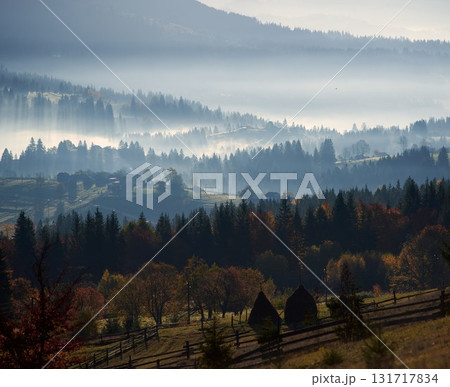 Morning sunlight streams through mist, illuminating serene mountain village nestled among forested hills. Light creates ethereal rays that gently touch autumn foliage and houses scattered on slopes. 131717834