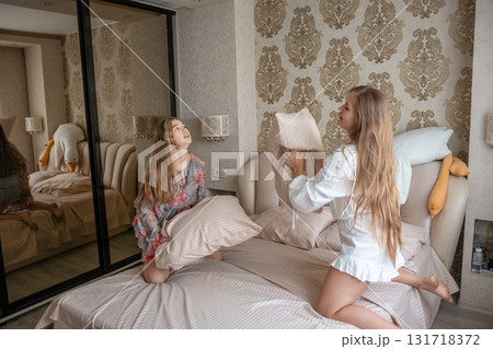 Pillow Fight Bedroom Girls Fun: Two young women playfully fight pillows on a bed inside a modern bedroom carefree enjoyment. 131718372