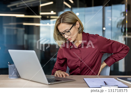 Businesswoman with eyeglasses experiencing back pain while working on laptop in a modern office, touching her back with hand and showing painful expression Businesswoman with eyeglasses experiencing back pain while working on laptop in a modern office, touching her back with hand and showing painful expression 131718575