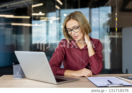 Stressed businesswoman is massaging her neck while working on a laptop in a modern office, experiencing discomfort and tension due to prolonged computer use 131718576