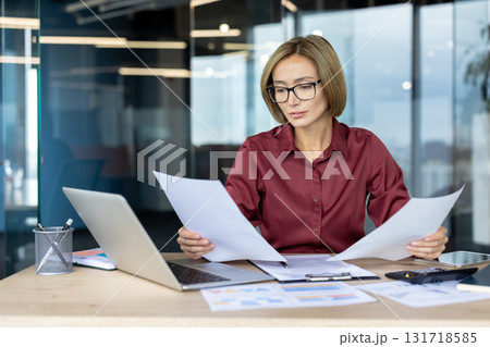 Focused businesswoman wearing glasses reads documents at a modern office desk, surrounded by a laptop, charts, and financial reports, exuding professionalism and concentration Focused businesswoman wearing glasses reads documents at a modern office desk, surrounded by a laptop, charts, and financial reports, exuding professionalism and concentration 131718585