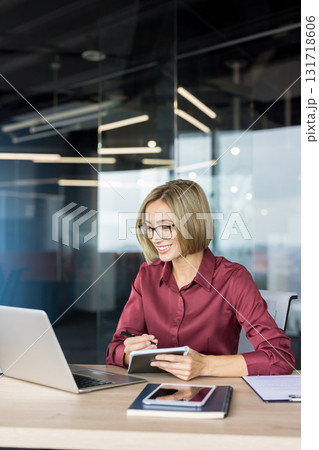 Smiling businesswoman in glasses taking notes while working on a laptop at her desk in a contemporary office, representing productivity and professional success Smiling businesswoman in glasses taking notes while working on a laptop at her desk in a contemporary office, representing productivity and professional success 131718606