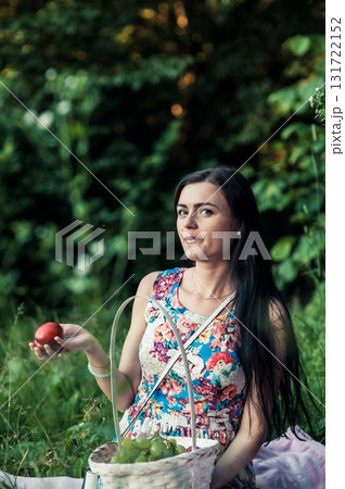 A young girl is sitting on a pink rug during a picnic, with a white basket of fruit standing nearby A young girl is sitting on a pink rug during a picnic, with a white basket of fruit standing nearby 131722152