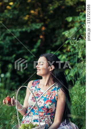 A girl in a floral dress sits at a picnic against a backdrop of green trees 131722166
