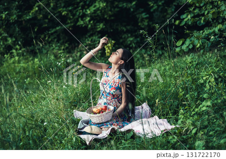 A lovely woman sits on a pink rug during a picnic, with a white basket of fruit standing nearby 131722170