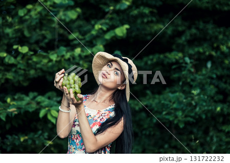 A lady in a hat with grapes in her hands stands on the grass. A young girl in a floral dress. A seductive girl against the backdrop of nature. 131722232