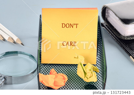 A notepad with dont care sits atop a wire mesh container holding crumpled paper near pencils and a magnifying glass, all on a light blue surface 131723443