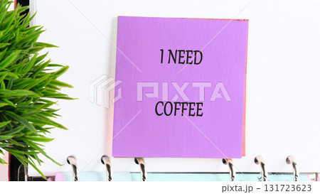 A purple sticky note with the words I NEED COFFEE is attached to a spiral notebook with a green artificial plant situated next to it, craving its morning or afternoon pick-me-up 131723623