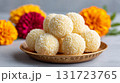 Festive ladoos made from condensed milk and rolled in coconut flakes are displayed on a traditional plate, surrounded by blooming marigold flowers 131723765