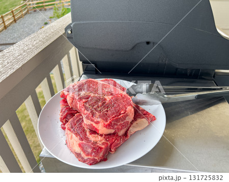 Two thick ribeye steaks sit on a plate with tongs, placed beside a grill outdoors. The steaks are ready for a summer barbecue under natural daylight. Two thick ribeye steaks sit on a plate with tongs, placed beside a grill outdoors. The steaks are ready for a summer barbecue under natural daylight. 131725832