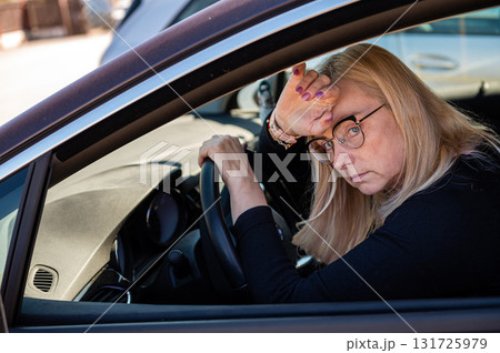 thoughtful middle-aged blonde in glasses sits behind the wheel of a car 131725979