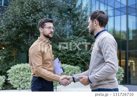 Two businessmen reaching an agreement, shaking hands outdoors near a modern office building, symbolizing success, trust, and a successful collaboration 131726710