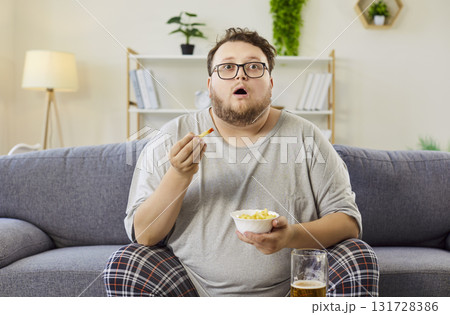 Overweight man eating fries on sofa, watching TV with beer at home, late evening Overweight man eating fries on sofa, watching TV with beer at home, late evening 131728386