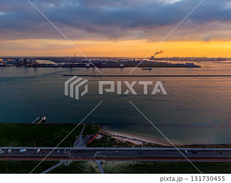 Aerial sunset view of coastal industrial zone road, beach, and grassy shoreline foreground contrast with smokestacks, cranes, and breakwater across golden water under dramatic cloudscape. 131730455
