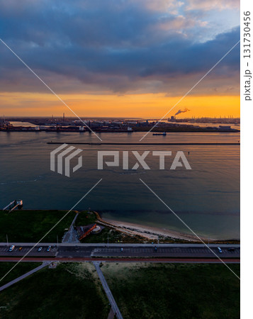 Aerial sunset view of coastal industrial zone road, beach, and grassy shoreline foreground contrast with smokestacks, cranes, and breakwater across golden water under dramatic cloudscape. 131730456