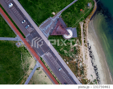Aerial view of coastal zone conical roofed pavilion near beach, road with parked cars, grassy fields, and clear water with depth gradient, blending geometric layout with natural shoreline. Aerial view of coastal zone conical roofed pavilion near beach, road with parked cars, grassy fields, and clear water with depth gradient, blending geometric layout with natural shoreline. 131730461