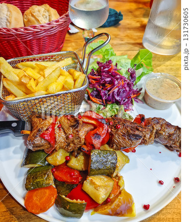 Close-up of a dinner plate with brochette bbq of grilled meat and pepper on a knife skewer served with roasted mediterranean vegetables and a basket of golden french fries, and sauce Close-up of a dinner plate with brochette bbq of grilled meat and pepper on a knife skewer served with roasted mediterranean vegetables and a basket of golden french fries, and sauce 131730605