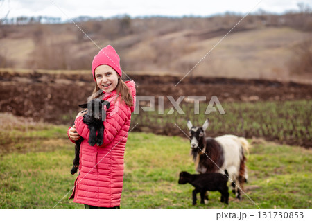Cute girl in red jacket and beanie holding black baby goat on farm. Rural lifestyle, childhood, animal care countryside setting Cute girl in red jacket and beanie holding black baby goat on farm. Rural lifestyle, childhood, animal care countryside setting 131730853