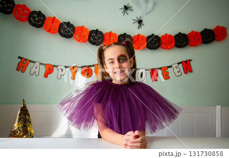 Smiling girl purple tulle costume with Halloween makeup, sitting decorated room with pumpkins, garlands, spiders, and witch hat. 131730858