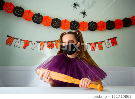 Girl dressed in purple costume with face makeup and black face mask holds wooden bat in room decorated for Halloween. Girl dressed in purple costume with face makeup and black face mask holds wooden bat in room decorated for Halloween. 131730862