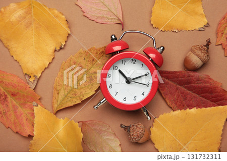 Red alarm clock, acorns and autumn leaves on brown background. Daylight saving time end Red alarm clock, acorns and autumn leaves on brown background. Daylight saving time end 131732311