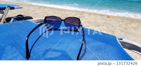 A pair of dark-framed sunglasses rests on a blue beach chair by the sea A pair of dark-framed sunglasses rests on a blue beach chair by the sea 131732426