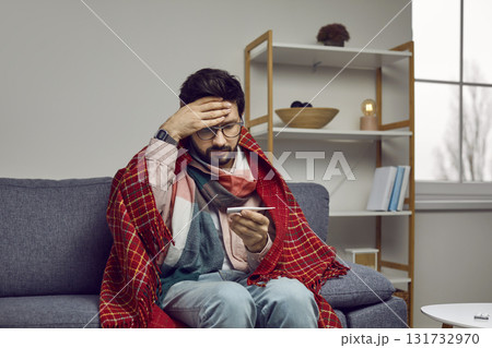 Sick man with fever looks confusedly at thermometer showing high body temperature. Sick man with fever looks confusedly at thermometer showing high body temperature. 131732970