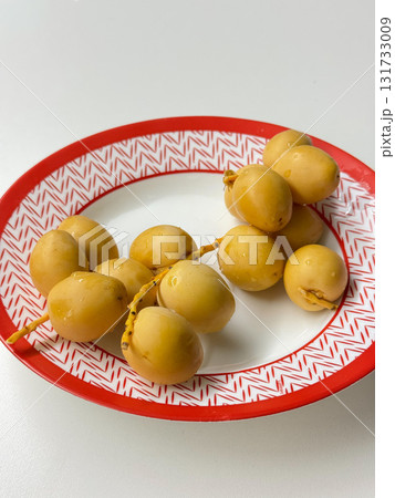 Fresh yellow dates with water drops lie on a white plate with a red geometric rim, illuminated by soft natural light 131733009