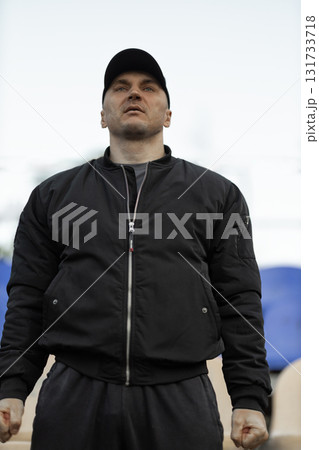 man in black bomber jacket and cap stands in empty stadium showing reactions to the team performance during an intense game feeling excitement and tension. 131733718