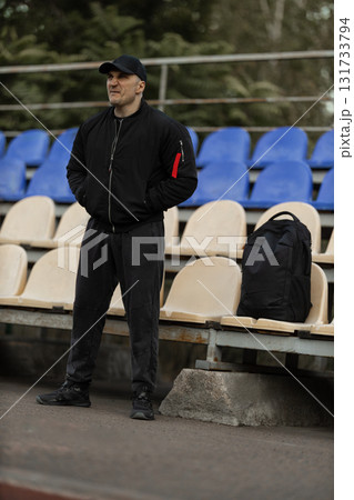 man stands in the empty stands wearing a black bomber jacket and cap focused on the unfolding sports game. His body language shows deep engagement with the match taking place. 131733794