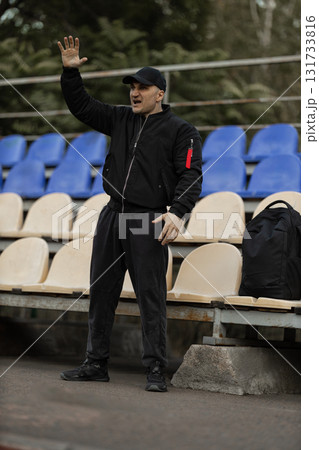 A man stands in the deserted stands of a stadium wearing a black bomber jacket and cap. He passionately reacts to the unfolding events during a team match showing intense emotions. A man stands in the deserted stands of a stadium wearing a black bomber jacket and cap. He passionately reacts to the unfolding events during a team match showing intense emotions. 131733816