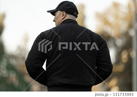 man wearing a black bomber jacket and sportswear stands on a treadmill in an outdoor setting. The environment features blurred trees in the background showing an active and healthy lifestyle. 131733993