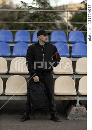 man wearing a black bomber jacket and cap stands among empty seats in a stadium tribune. He holds a backpack and gazes around seemingly lost in thought. man wearing a black bomber jacket and cap stands among empty seats in a stadium tribune. He holds a backpack and gazes around seemingly lost in thought. 131734057