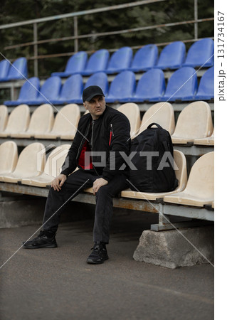 man wearing a black bomber jacket and cap sits on a bench in an empty stadium tribune. He has a backpack beside him and appears to be in a reflective mood in a quiet setting. 131734167