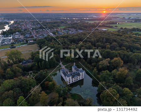 Aerial sunset view of historic castle with moat, nestled in dense forest and autumn foliage, bordered by residential zone, open fields, and distant river under glowing orange sky. Aerial sunset view of historic castle with moat, nestled in dense forest and autumn foliage, bordered by residential zone, open fields, and distant river under glowing orange sky. 131734492