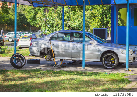 Description: Silver sedan lifted on a car jack for tire replacement in an outdoor auto repair area. Concept of car maintenance, repair work in progress, change of seasons and wheels Description: Silver sedan lifted on a car jack for tire replacement in an outdoor auto repair area. Concept of car maintenance, repair work in progress, change of seasons and wheels 131735099