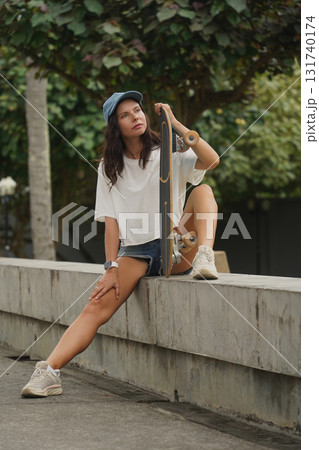 A young woman sits comfortably on a low wall at the park holding a skateboard in her hands A young woman sits comfortably on a low wall at the park holding a skateboard in her hands 131740174