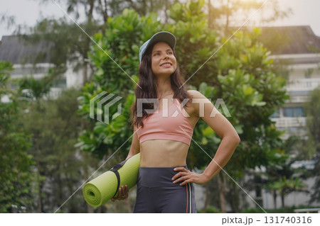 A young woman happily prepares to do yoga on a mat in a beautiful park. 131740316