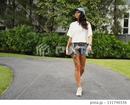 A woman is walking at the city park while carrying a skateboard in her hands A woman is walking at the city park while carrying a skateboard in her hands 131740336
