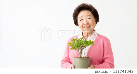 観葉植物の鉢を手に持ち、歯を見せて明るく微笑むピンクのカーディガンを着たシニア女性。 観葉植物の鉢を手に持ち、歯を見せて明るく微笑むピンクのカーディガンを着たシニア女性。 131741342
