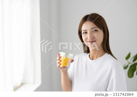 光が差し込む明るい窓辺でオレンジジュースが入ったグラスを持つ若い女性 131742064